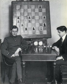 Capablanca y Alekhine posando antes de una partida, con un tablero mural gigante en la pared