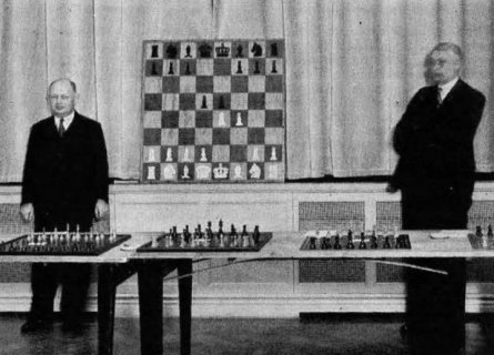 Spielmann de pie frente a un tablero mural que contiene las dos primeras jugadas de un gambito de rey: 1.e4 e5 2.f4 d5. Delante de l tiene 3 juego de ajedrez y a su derecha aparece el organizador del evento.